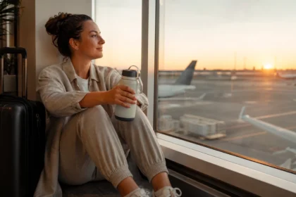 Woman sitting at airport window in sunlight with water bottle, preparing for a long-haul flight naturally