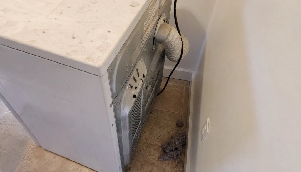 Back of a residential dryer showing a flexible silver vent duct with lint buildup near the base